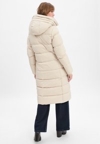 Beige gewatteerde jas met een hoge kraag, voorzien van een capuchon en horizontaal gestikte structuur. Draagt een combinatie van donkere wijduitlopende jeans en zwarte laarzen.