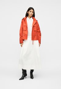 Femme portant une veste à capuche orange vif sur une chemise blanche à boutons et une longue jupe blanche avec des bottes noires à talons pointus.