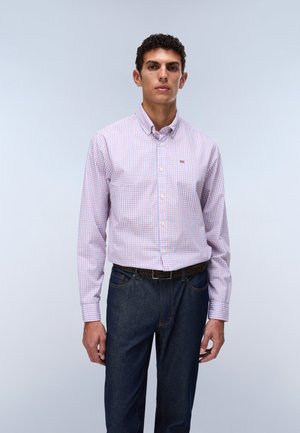 Ung mand iført en langærmet, pink og blå ternet skjorte, der er stoppet ned i mørkeblå jeans med et sort bælte, stående mod en lys baggrund.