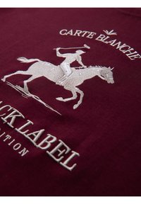 Broderie d'un joueur de polo à cheval avec un maillet levé, au-dessus le texte "CARTE BLANCHE" et "BLACK LABEL" sur un tissu bordeaux.