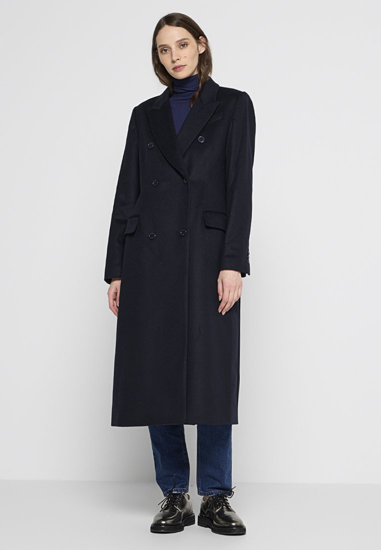 Donker marineblauwe dubbelbreasted jas met een lange silhouet, op maat ontworpen, met geknipte revers en zijzakken, gedragen over een blauwe coltrui.