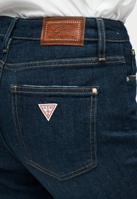 Σκούρα μπλε τζιν από τζιν ύφασμα με δερμάτινο ταμπελάκι που φέρει ανάγλυφο το λογότυπο "Guess", λευκό τριγωνικό λογότυπο στην πίσω δεξιά τσέπη και αντίθετη ραφή.