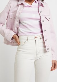 Kobieta nosząca jasnoróżową dżinsową kurtkę na różowo-białej koszulce polo w paski, do której dobrała wysokie białe dżinsy.