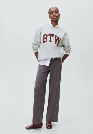 Persona con felpa grigia con la scritta "BTW", camicia bianca sotto, pantaloni larghi marrone, e mocassini bordeaux, in piedi con le mani dietro la schiena.