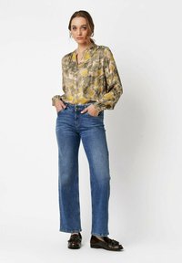 Frau trägt eine blütenartige Bluse in Gelb und Grün, blaue gerade Jeans und schwarze Nieten-Lederschuhe, steht mit den Händen in den Taschen.