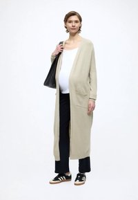 Długi beżowy kardigan z przednimi kieszeniami, noszony na białej bluzce i czarnych spodniach. Czarne trampki z białymi paskami oraz duża czarna torba typu tote.