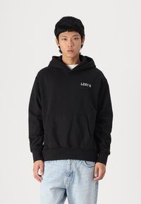 Felpa nera in cotone, dotata di tasca a marsupio, polsini a costine e logo "Levi's" bianco sul petto. Indossata con jeans azzurri chiari.
