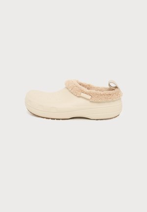 Chaussure beige sans lacets avec un revers doux et duveteux et une semelle texturée, conçue pour un usage décontracté en intérieur ou en extérieur.
