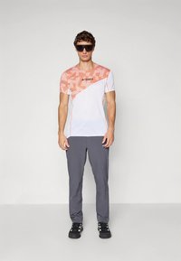 Lettvekts t-skjorte i stoff med et diagonalt stripemønster i gradient fra oransje til hvitt. Kombinert med grå treningsbukser og svarte sko.