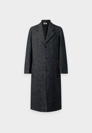 Jeune homme portant un long manteau texturé foncé, une chemise blanche, un pantalon sombre et des mocassins noirs, debout avec une main dans la poche.