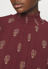 Gros plan d’une personne à la peau foncée portant un chemisier bordeaux avec un col montant à volants et de petits motifs beiges semblables à des paisleys.
