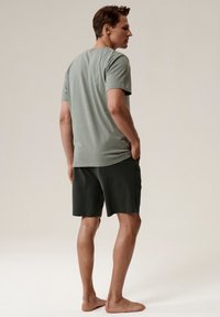 Jasnozielona koszulka z krótkim rękawem z miękkiego materiału, w połączeniu z ciemnozielonymi szortami. Model stoi boso, ręce w kieszeniach, skierowany bokiem.