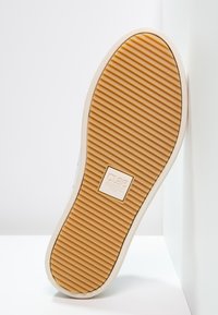 Semelle de chaussure avec un motif texturé à rayures en caoutchouc beige. Présente un bord blanc et un logo de marque en relief au centre.