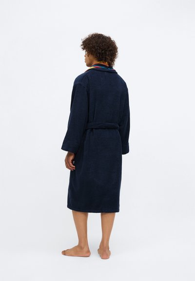 Kortsirged juuksed inimene, kes kannab tumesinist hommikumantlit erksate kraega, seiseb paljajalu, seljaga kaamera poole, valge taustaga.