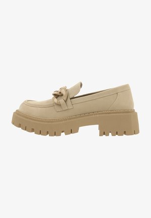 Mocassin en daim beige avec une semelle en caoutchouc épaisse et texturée, orné d'un nœud décoratif à l'avant et de détails de surpiqûres.