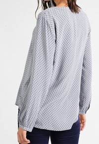 Pitkähihaisessa vaaleansinisessä paidassa on toistuva tummansininen kuosi. Siinä on rypytetty olkapääosuus ja joustavat hihansuut mukavan istuvuuden takaamiseksi.