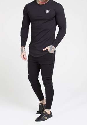 Hombre vestido con un conjunto ajustado completamente negro, con camisa de manga larga, pantalones ajustados y zapatillas negras con detalles dorados, sobre un fondo blanco liso.