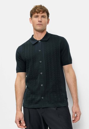 Homme portant une chemise en maille noire à manches courtes avec col et boutons, posant devant un fond clair uni.