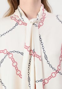 Camicia bianca con motivo a catena rosso e navy, caratterizzata da un dettaglio di scollatura legata e una texture liscia.