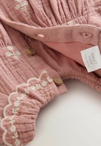 Light pink fabric bodysuit with embroidered floral details, wooden buttons, and gathered texture along the neckline and cuffs. Tag visible.