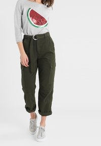 Maglia grigia a maniche lunghe con un design di anguria rossa e verde, abbinata a pantaloni cargo verde oliva e sneakers grigie.
