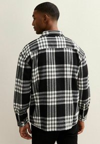 Uomo di spalle che indossa una camicia a maniche lunghe a scacchi bianco e nero con polsini abbottonati e pantaloni neri.