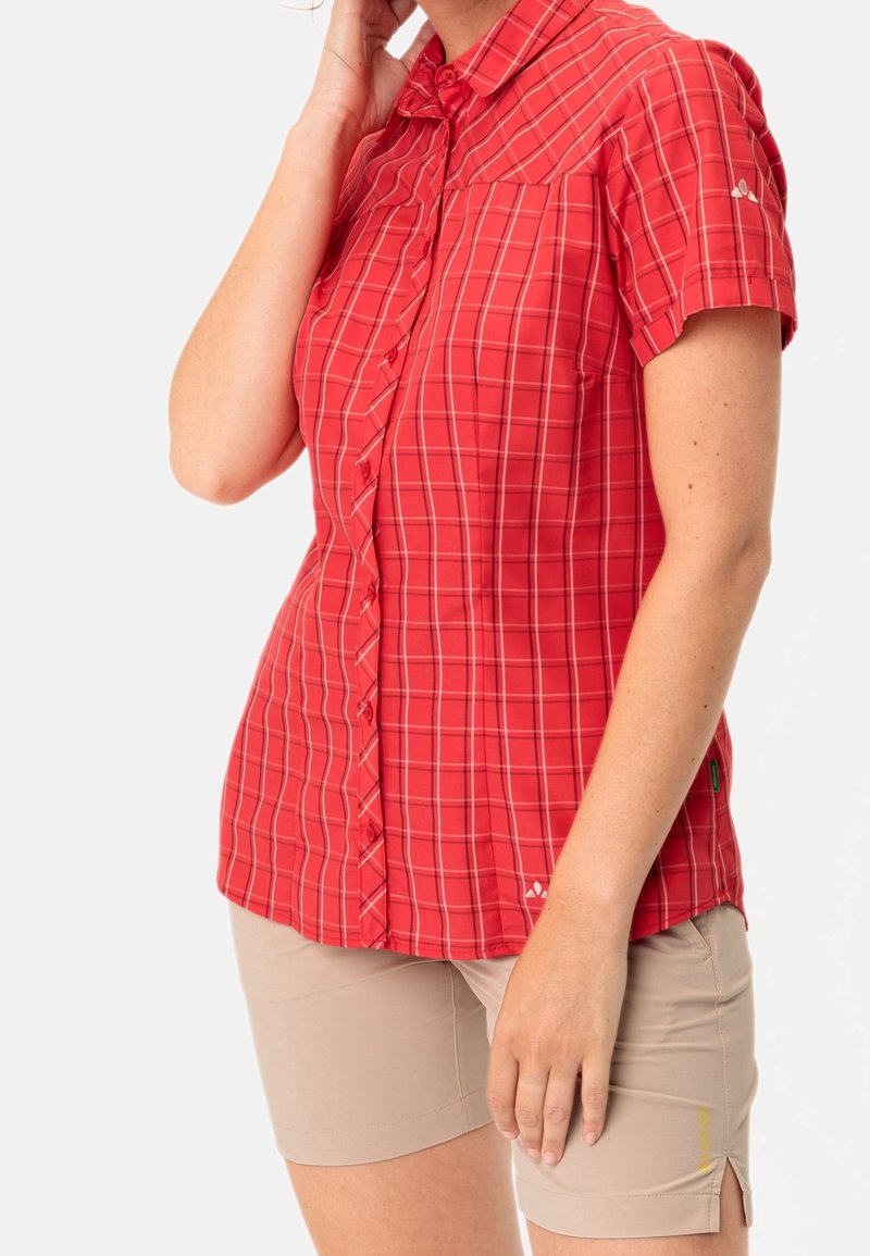 Camicia a maniche corte a scacchi rossi con colletto, chiusura a bottoni e un accento con logo. Abbinata a pantaloni corti beige chiaro, che mostrano un design casual.
