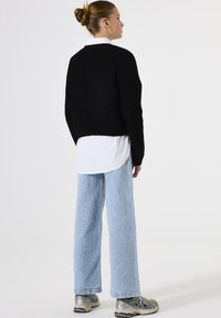 Vrouw met een haar in een bun, gekleed in een zwarte trui over een wit shirt, lichtblauwe wijde jeans en zilveren sneakers, zijwaarts gekeerd.