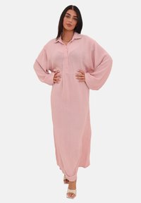 Donna che indossa un lungo vestito rosa con bottoni e grandi maniche, in piedi con le mani sui fianchi e con sandali con tacco leggeri.
