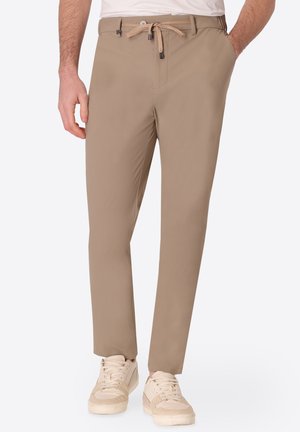 Mann trägt beige Kordelzughose mit Seitentaschen und hellbeige Sneakers, steht mit einer Hand in der Tasche.