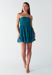 Turquoisekleurige strapless jurk met een geplooide bodice en een gelaagde rok. Lichtgewicht stof met een vloeiende textuur, aangevuld met strappy gouden sandalen.