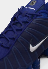 Gros plan d'une basket Nike Shox bleue avec une tige texturée, un Swoosh Nike blanc et un amorti Shox distinct sous la semelle.