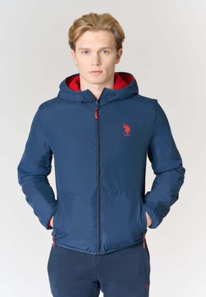 Giacca con cappuccio blu navy con dettagli rossi, dotata di chiusura con zip, tasche laterali e logo sul petto. Texture liscia, materiale leggero.