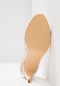 Chaussure à talon haut beige pour femme à bout pointu avec dessus en dentelle, vue de la semelle lisse couleur cuir clair sur fond blanc d'angle.