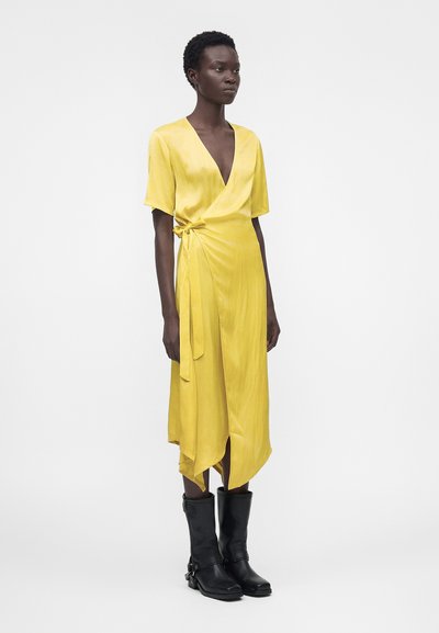 Γυναίκα στέκεται φορώντας κίτρινο φόρεμα τύπου wrap με κοντά μανίκια και μαύρες μπότες μέχρι τη μέση της γάμπας σε απλό λευκό φόντο.