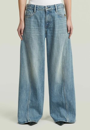 Jean bleu clair à jambes larges porté par une personne debout, les bras détendus le long du corps, associé à des chaussures noires pointues visibles.