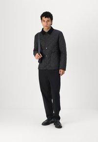 Casaco preto acolchoado com botões de pressão, colarinho em contraste e bolsos frontais. Usado com calças e sapatos pretos, o modelo está posicionado contra um fundo branco.