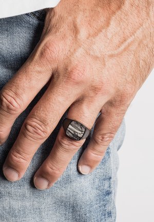 Schwarzer Ring mit einem quadratischen Stein, der ein marmoriertes Grau- und Weißmuster aufweist. Am Handgelenk über einem Denim-Stoff getragen. Texturierte Haut sichtbar.
