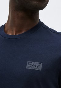 Άνδρας που φοράει μπλε ναυτικό t-shirt με στρογγυλή λαιμόκοψη και μικρό λογότυπο EA7 Emporio Armani στο αριστερό στήθος, μπροστά σε μια απλή φόντο.