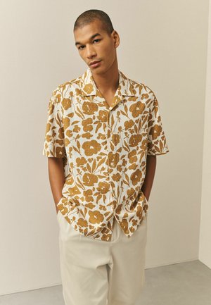 Mladý muž s krátkymi vlasmi, oblečený v krátkoročnom košeli s horčicovým kvetinovým vzorom a svetlo béžových nohaviciach, ruky v vreckách.