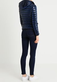 Navy puffer jacket with a hood, featuring a quilted design and glossy finish. Paired with dark denim skinny jeans and white sneakers.