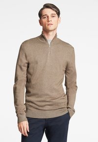 Tan zip-neck genser laget av strikket stoff, med lange ermer og ribbestrikkede mansjetter. Subtil tekstur, slim fit, parret med mørke bukser.