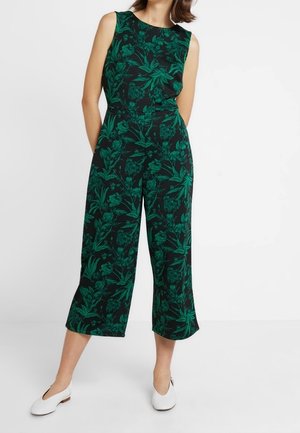 Femme portant une combinaison noire sans manches avec un imprimé floral vert et des chaussures plates blanches, debout les mains dans le dos.