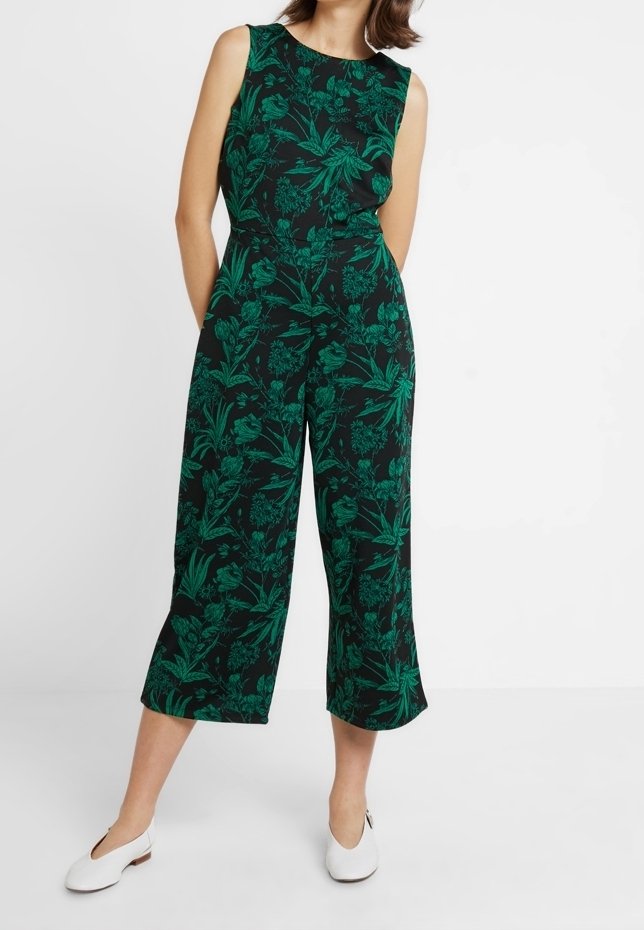 Femme portant une combinaison noire sans manches avec un imprimé floral vert et des chaussures plates blanches, debout les mains dans le dos.