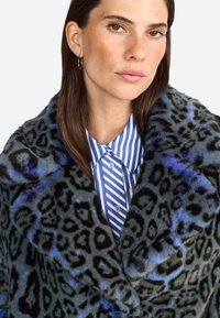 Manteau en fausse fourrure présentant un imprimé léopard bleu et noir, avec une texture douce. En dessous, une chemise rayée bleu et blanc est visible.