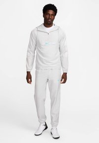 Lichtgrijze Nike capuchonjack met een ritszak op de voorkant en een turquoise logo, gecombineerd met bijpassende joggerbroeken en witte schoenen.