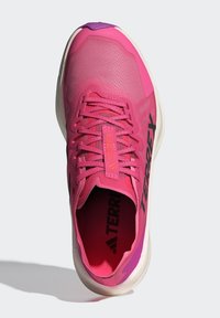 Růžová sportovní bota s texturovaným síťovinovým svrškem, plochými tkaničkami a kontrastním černým logem. Má polstrovanou podrážku a šikmou design paty.