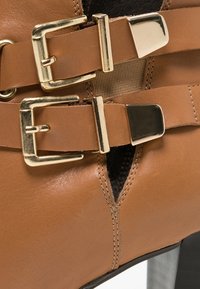 Bottine en cuir tan avec deux boucles et sangles dorées au-dessus d'un talon épais noir.