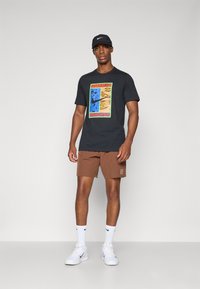 Svart t-skjorte med fargerik grafisk design, brune shorts, hvite sokker og hvite joggesko. Alle klær har sporty stil.