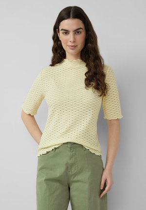 Vrouw met lang donker haar, gekleed in een lichtgeel, gestructureerd, kort mouwachtig shirt en groene, hooggetailleerde broek, staand tegen een grijze achtergrond.